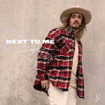 Jordan Feliz wearing a checkered clothing and a hat while standing near a wall, cover art for his song "Next To Me" - My Christian Musician