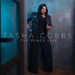 Tasha Cobbs Leonard poses for a photo wearing a black leather jacket over a black jumpsuit, with a text overlay that displays "TASHA COBBS ONE PLACE LIVE" - My Christian Musician